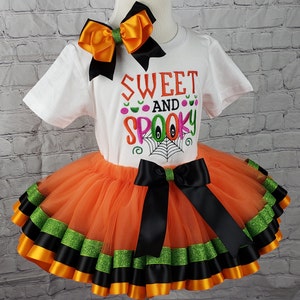 K&ouml;nnte beinhalten: Ein wei&szlig;es T-Shirt mit dem Text "Sweet and Spooky" in bunter Stickerei. Das Shirt wird mit einem orangefarbenen T&uuml;ll-Tutu mit schwarzem und gr&uuml;nem Bandbesatz kombiniert. Eine passende orange-schwarze Schleife ist im Lieferumfang enthalten.