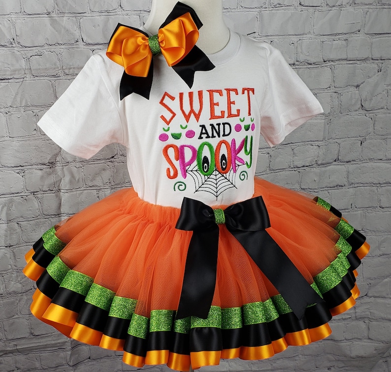 K&ouml;nnte beinhalten: Ein wei&szlig;es T-Shirt mit dem Text "Sweet and Spooky" in bunter Stickerei. Das Shirt wird mit einem orangefarbenen T&uuml;ll-Tutu mit schwarzem und gr&uuml;nem Bandbesatz kombiniert. Eine passende orange-schwarze Schleife ist im Lieferumfang enthalten.