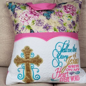 May include: A white pillow with a pink floral fabric on the back. The front of the pillow has a gold glitter cross with turquoise embroidery. The text "Tell me the story of Jesus Write on my heart every word" is embroidered in turquoise and pink.