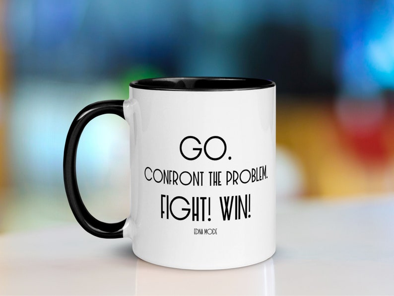 May include: White ceramic coffee mug with a black rim and handle. The mug has a black inspirational quote that reads "GO. CONFRONT THE PROBLEM. FIGHT! WIN! EDNA MODE".