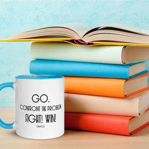 May include: A white coffee mug with a blue rim and handle. The mug has black text that reads "GO. CONFRONT THE PROBLEM. FIGHT! WIN!  -  EDM MCBride". The mug is sitting on a wooden surface in front of a stack of colorful books.