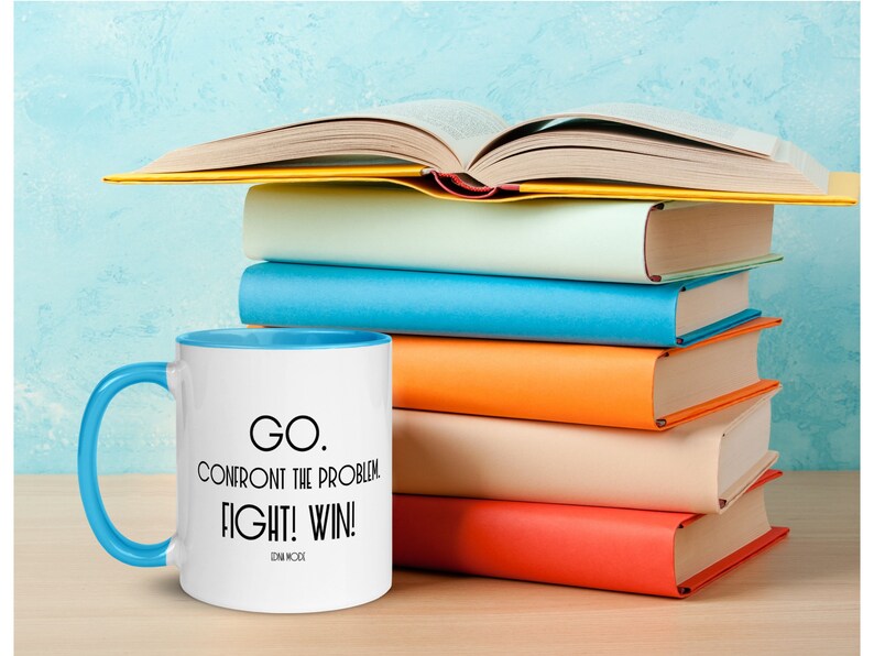 May include: A white coffee mug with a blue rim and handle. The mug has black text that reads "GO. CONFRONT THE PROBLEM. FIGHT! WIN!  -  EDM MCBride". The mug is sitting on a wooden surface in front of a stack of colorful books.