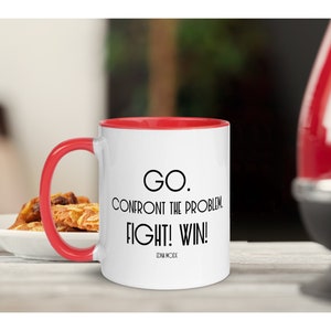 May include: White ceramic coffee mug with a red rim. The mug has a black quote that reads "GO. CONFRONT THE PROBLEM. FIGHT! WIN!" with the name "DAN WOOD" at the bottom.