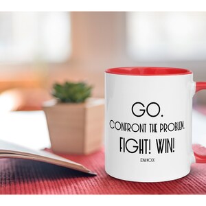 May include: White ceramic mug with a red rim and handle. The mug has a black motivational quote that reads "GO. CONFRONT THE PROBLEM. FIGHT! WIN!" with the words "EDNA MODE" printed below the quote.