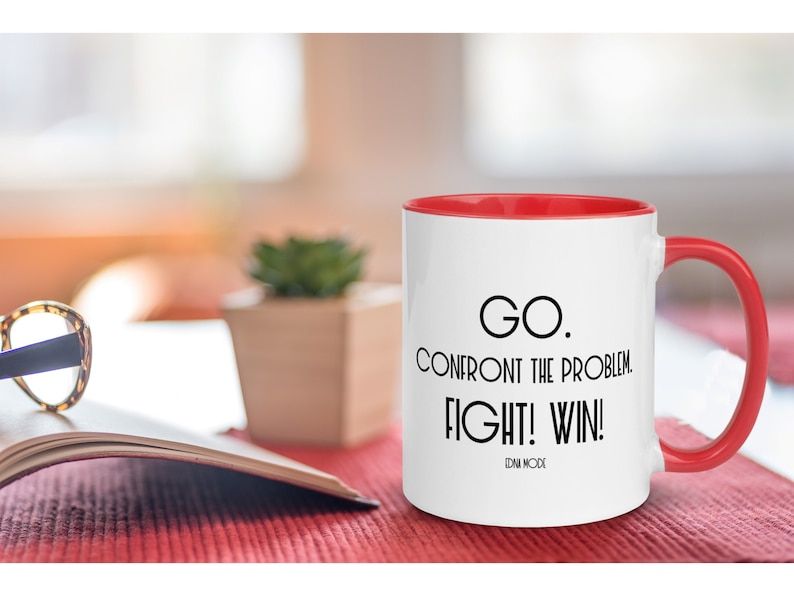 May include: White ceramic mug with a red rim and handle. The mug has a black motivational quote that reads "GO. CONFRONT THE PROBLEM. FIGHT! WIN!" with the words "EDNA MODE" printed below the quote.