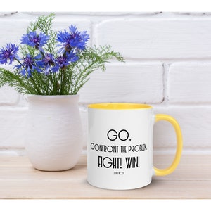 May include: White ceramic mug with a yellow rim and handle. The mug has black text that reads "GO. CONFRONT THE PROBLEM. FIGHT! WIN!" with the words "DANA MCKEE" in smaller font below.