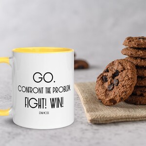 May include: A white ceramic mug with a yellow rim and handle. The mug has black text that reads "GO. CONFRONT THE PROBLEM. FIGHT! WIN! EDNA MODE".