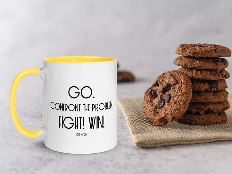 May include: A white ceramic mug with a yellow rim and handle. The mug has black text that reads "GO. CONFRONT THE PROBLEM. FIGHT! WIN! EDNA MODE".