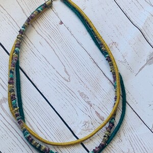 May include: A multi-strand necklace made of woven fabric in shades of mustard yellow, forest green, and burgundy. The necklace has a simple metal clasp.