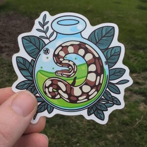 May include: A sticker featuring a brown and white snake coiled inside a blue glass bottle with green liquid. The bottle is surrounded by green leaves and a light blue border. The sticker is held by a hand.