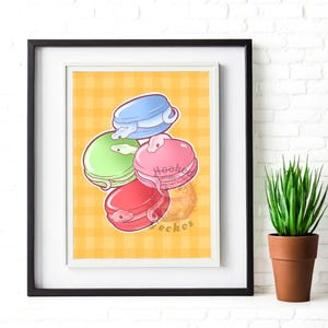 以下が含まれることがあります： 4色のマカロンの気まぐれなイラストが描かれた額入りのプリント。マカロンは青、緑、ピンク、赤で、それぞれヘビのデザインが施されています。背景は黄色のチェック柄です。