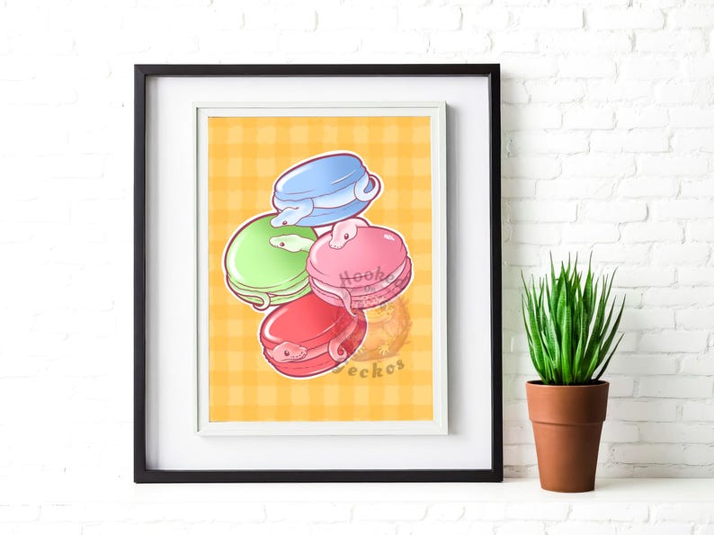 以下が含まれることがあります： 4色のマカロンの気まぐれなイラストが描かれた額入りのプリント。マカロンは青、緑、ピンク、赤で、それぞれヘビのデザインが施されています。背景は黄色のチェック柄です。