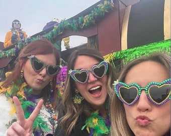 May include: Three women wearing heart-shaped sunglasses with rhinestones and beads. The sunglasses are purple, green and black. The women are wearing Mardi Gras beads and feathers.