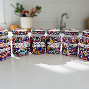 Op de afbeelding: Zes decoratieve glazen potten bedekt met kleurrijke strass-steentjes en kralen. Elke pot heeft een wit deksel en een label met een ander woord gespeld in witte letterkralen: HAPPY PILLS, ZOLOOF QUEEN, HEART PILLS, CHILL PILLS, DERALLE en XAPRO.