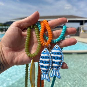 May include: A collection of colorful beaded necklaces with fish pendants. The necklaces feature beads in green, orange, and turquoise, with fish pendants in blue and white. The fish pendants have a whimsical design, adding a playful touch to the jewelry.