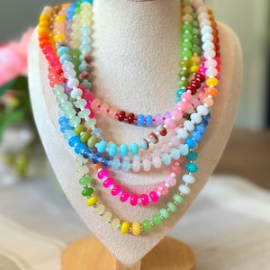 May include: A display of colorful beaded necklaces. The necklaces are arranged in layers, showcasing a spectrum of colors including pink, green, yellow, blue, and orange. Each necklace features small, round beads and silver-toned clasps.