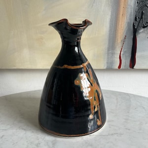 Puede incluir: Un jarrón de cerámica negro con un esmalte marrón y una parte superior rizada. El jarrón tiene un diseño único y abstracto con acentos marrones y naranjas.