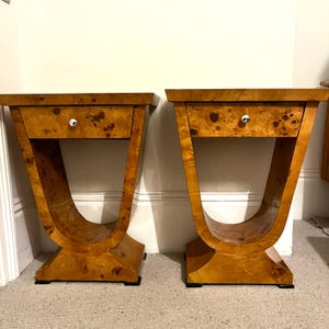 May include: Pair of Art Deco style bedside tables. Each table features a drawer with a round knob and a curved, pedestal base. The tables are made of wood with a warm, light brown finish and a unique wood grain pattern.