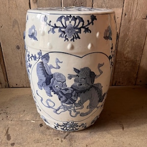 May include: A barrel-shaped ceramic garden stool featuring a cream-coloured glaze with intricate blue hand-painted illustrations of two guardian lions playing with a ball, accented by floral motifs and a row of raised circular studs along the top and base.