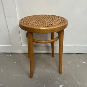 May include: A round, light-brown wooden stool with a woven cane seat. The stool has four legs and a curved support piece. The wood has a natural finish, and the stool appears to be vintage or antique.