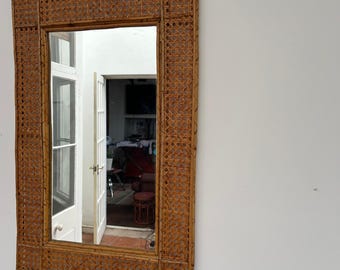 May include: A rectangular mirror with a woven rattan frame. The frame is a light brown colour and has a geometric pattern. The mirror is reflecting a room with a door and a chair.