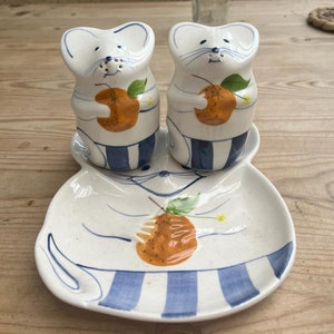 以下が含まれることがあります： オレンジを持ったネズミの形をした白いセラミック製の塩とコショウのセット。シェーカーは、青と白のストライプのデザインが施された白いセラミック製の皿の上に置かれています。