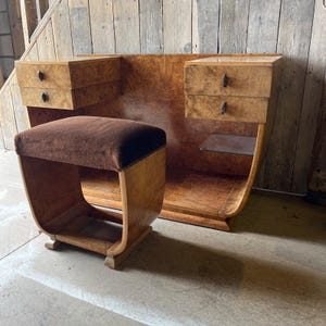 Puede incluir: Un tocador vintage con un taburete. El conjunto presenta una gran parte trasera de madera curvada con dos pequeños cajones a cada lado. El taburete tiene un asiento de terciopelo marrón y un marco de madera curvado. La madera tiene un acabado cálido y natural.