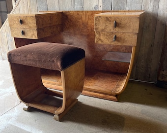 May include: A vintage vanity set with a stool. The set features a large, curved wooden back with two small drawers on each side. The stool has a brown velvet seat and a curved wooden frame. The wood has a warm, natural finish.