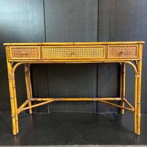 May include: A bamboo desk with three drawers. The drawers have woven cane fronts and small, round knobs. The desk has a natural, light brown finish and a sturdy, open frame design. The legs are curved and connected by horizontal supports.
