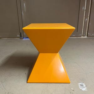 May include: A bright orange, hourglass-shaped side table with a square top.