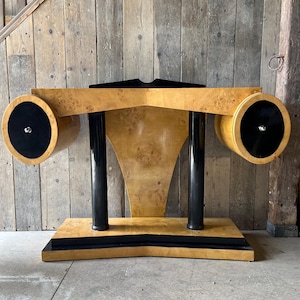 Puede incluir: Un escritorio de estilo Art Déco con un acabado de veta de madera clara. El escritorio presenta dos elementos cilíndricos con acentos circulares negros en cada extremo. El escritorio tiene soportes verticales negros y una base negra.