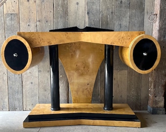 May include: An Art Deco-style desk with a light wood grain finish. The desk features two cylindrical elements with black circular accents on each end. The desk has black vertical supports and a black base.