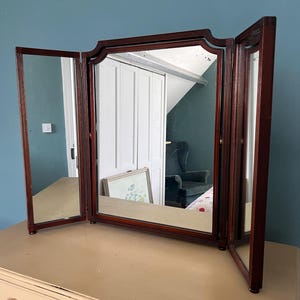 May include: A tri-fold vanity mirror with a dark wood frame featuring vertical fluted detailing. The central rectangular mirror is flanked by two adjustable side panels, all mounted on a base, providing a wide reflective surface area.