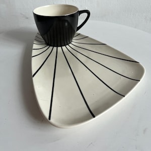 Puede incluir: Un juego de taza y platillo de cerámica en blanco y negro. El platillo es triangular con líneas negras que irradian desde el centro. La taza es negra con líneas negras verticales.