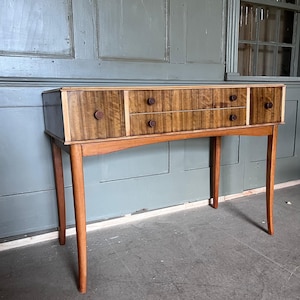 Puede incluir: Una consola de madera de mediados de siglo con tres cajones y un estante inferior. La mesa tiene un acabado de veta de madera cálida con perillas marrones oscuras y patas delgadas y cónicas. La pieza está colocada contra una puerta verde apagada.