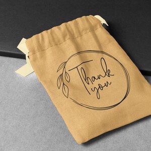 May include: A small, brown fabric drawstring bag with a black circle design that says "Thank you" in cursive. The bag is on a grey surface.