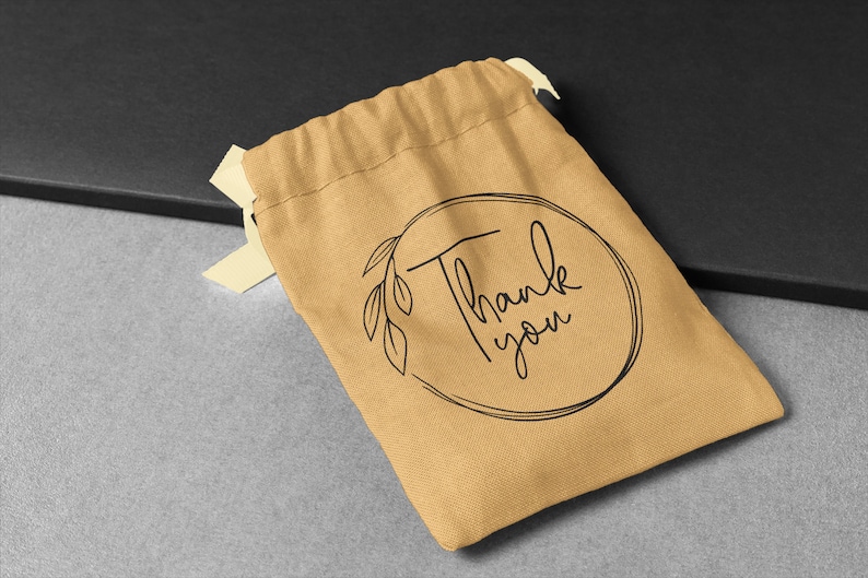 May include: A small, brown fabric drawstring bag with a black circle design that says "Thank you" in cursive. The bag is on a grey surface.