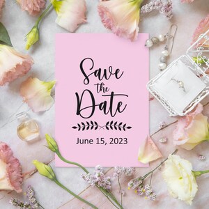 May include: Pink save the date card with black calligraphy that reads "Save the Date" and "June 15, 2023". The card is surrounded by pink and white flowers.
