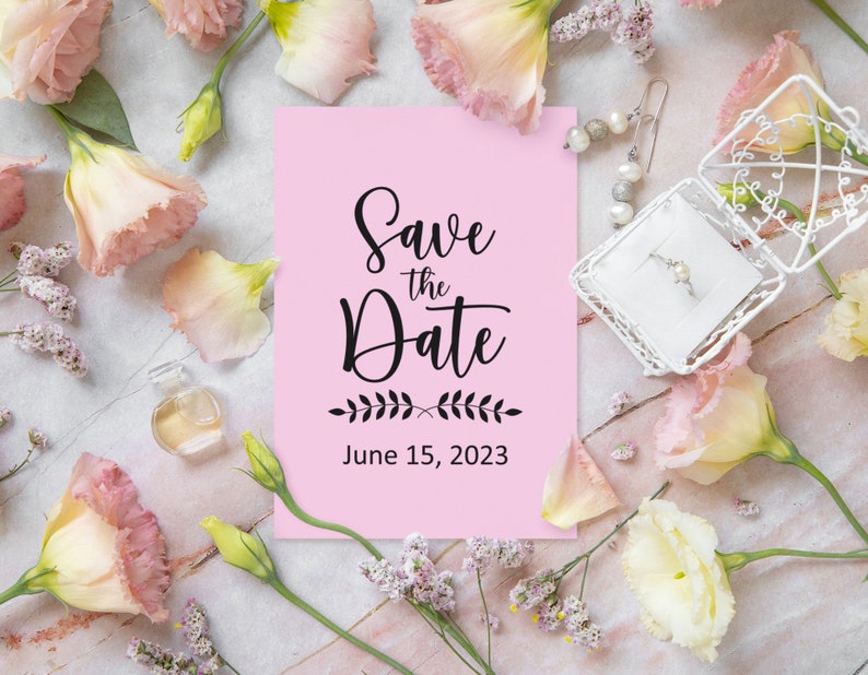 May include: Pink save the date card with black calligraphy that reads "Save the Date" and "June 15, 2023". The card is surrounded by pink and white flowers.
