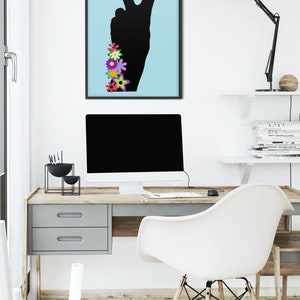 May include: A framed print of a black peace sign silhouette with colorful flowers on a light blue background. The print is hanging on a white wall above a desk with a computer, a lamp, and a chair.
