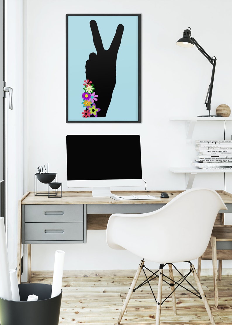 May include: A framed print of a black peace sign silhouette with colorful flowers on a light blue background. The print is hanging on a white wall above a desk with a computer, a lamp, and a chair.