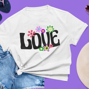 May include: White t-shirt with a black "LOVE" graphic in a retro font. The graphic is surrounded by colorful flowers with pink, purple, green, and yellow petals.