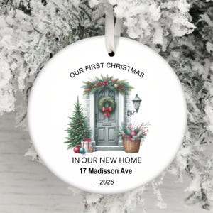 May include: A round white ceramic Christmas ornament with a white ribbon hanger. The design features a gray front door with a wreath, a Christmas tree, gifts, and a lantern. Text reads: OUR FIRST CHRISTMAS IN OUR NEW HOME 17 Madisson Ave - 2026 -.