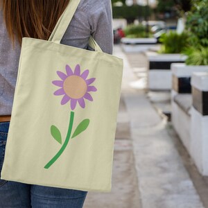 May include: A person carrying a light yellow tote bag with a purple flower design. The flower has a yellow centre and green leaves.