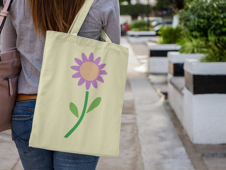 May include: A person carrying a light yellow tote bag with a purple flower design. The flower has a yellow centre and green leaves.