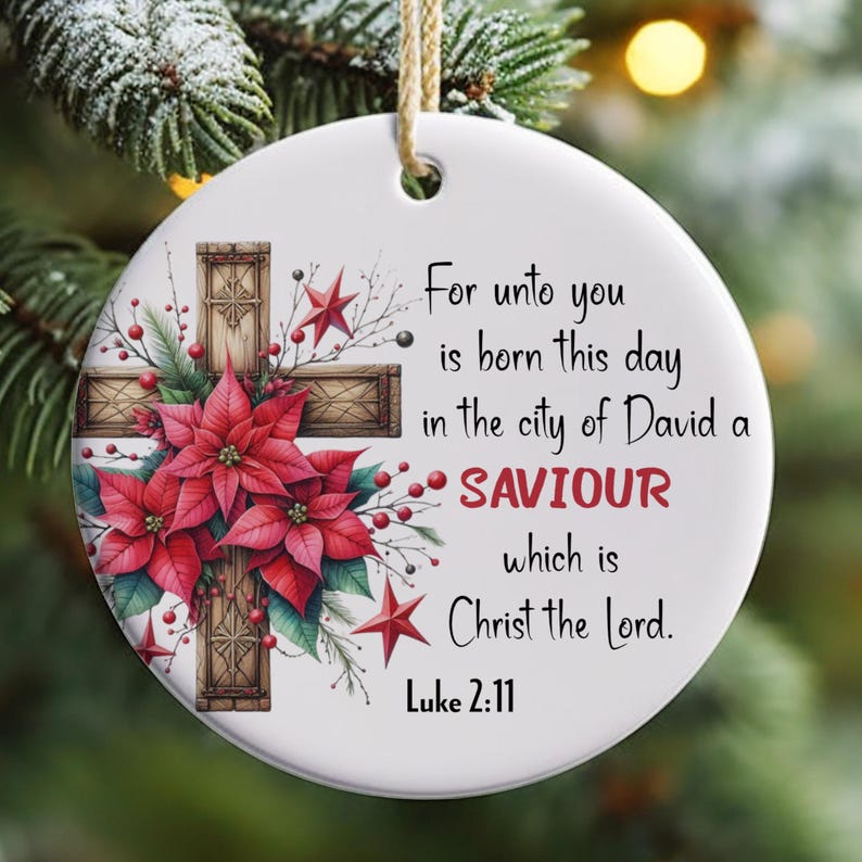 May include: A white round Christmas bauble featuring a red poinsettia and a wooden cross. The bauble has the text "For unto you is born this day in the city of David a SAVIOUR which is Christ the Lord. Luke 2:11"
