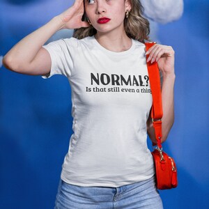 May include: A woman wearing a white t-shirt with the text "NORMAL? Is that still even a thing" in black lettering. She is wearing blue jeans and a red crossbody bag.