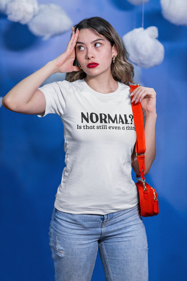 May include: A woman wearing a white t-shirt with the text "NORMAL? Is that still even a thing" in black lettering. She is wearing blue jeans and a red crossbody bag.