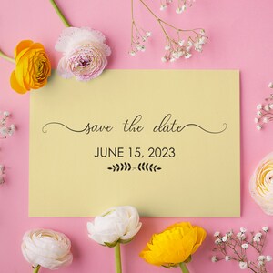 May include: A yellow and white save the date card with the text "save the date" and "JUNE 15, 2023" surrounded by white and yellow flowers on a pink background.
