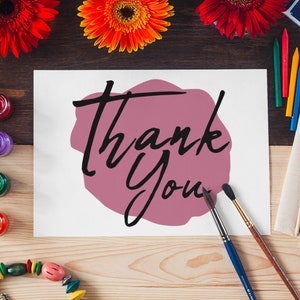 May include: A white card with the words "Thank You" written in black script on a pink watercolour background. The card is surrounded by paintbrushes, paint, and flowers.